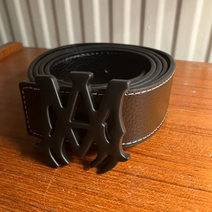 Amiri bälte - Säljer ett svart Amiri bälte med svart buckle, i grovpräglat skinn med vit kontrastsöm längs kanterna. Bältet har ett stort, matt Amiri spänne som verkligen sticker ut och ger en cool och lyxig vibe. Perfekt för dig som vill ha en streatweare-accessoar till ett par fina jeans. Nytt skick!