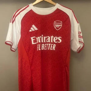Arsenal hemmatröja Adidas röd/vit L - Arsenal 25/26 hemmatröja från Adidas i storlek L. Röd bas med vita ärmar och detaljer, klassiskt klubbmärke och Adidas-logga på bröstet. Snyggt mönster över hela framsidan och tryckta sponsorloggor. Tillverkad i lätt och ventilerande polyester. Ny pris 1199
