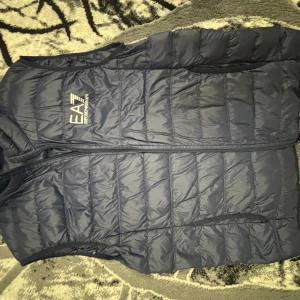 Mörkblå dunväst från EA7 Emporio Armani - Snygg mörkblå dunväst från EA7 Emporio Armani med quiltad design och dragkedja framtill. Västen har en stilren look med logga på bröstet och är perfekt för lager-på-lager-stil. Tillverkad i lätt och värmande syntetmaterial.
