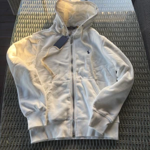 Vit hoodie från Polo Ralph Lauren - Snygg vit hoodie från Polo Ralph Lauren med klassisk blå logga på bröstet. Tröjan har huva med snörning, dragkedja framtill och två fickor. Insidan är mjuk och fleecefodrad för extra komfort. Perfekt för chill dagar och streetwear-stil.