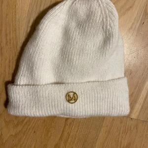 Vit stickad mössa med gulddetalj - Snygg vit stickad mössa med uppvikt kant och en liten guldfärgad M-logga framtill. Perfekt för kalla dagar och enkel att matcha med din vinterstil. Mjuk och skön passform som håller dig varm och ser clean ut.