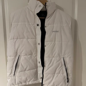 Vit dunväst från Peak Performance VINTAGE - Snygg vit dunväst från Peak Performance med quiltad design och hög krage. Västen har två fickor med dragkedja och stängs med både dragkedja och tryckknappar. Perfekt för lager-på-lager och chillare dagar. 