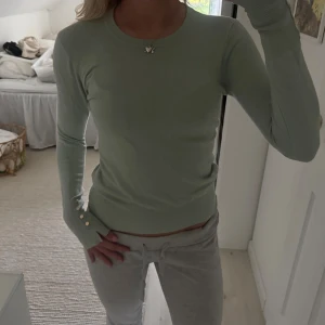 Ljusgrön stickad tröja med knappar - Supersnygg ljusgrön stickad tröja med rund hals och långa ärmar. Tröjan har dekorativa guldknappar vid ärmsluten och en tight passform som sitter riktigt snyggt. Perfekt att styla med jeans eller kjol för en fräsch look.