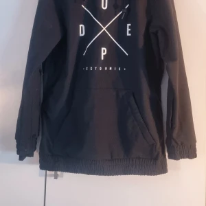 Svart snowboardjacka Yeti W - Svart snowboardjacka från Dope med stor magficka, elastiska muddar och tryckt grafisk logga framtill. Jackan har huva med snörning och är tillverkad i slitstarkt syntetmaterial, perfekt för vinterns äventyr i backen. Använd vid ett tillfälle.