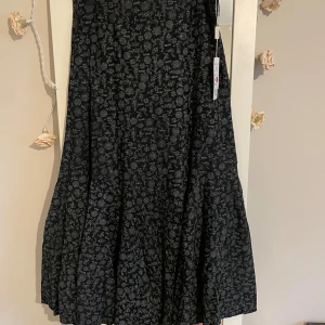 Svart långkjol med blommigt mönster YILING - Svart långkjol  med grått blommigt mönster. Kjolen har en vid och flowy passform med volang nedtill, vilket ger en snygg siluett. Perfekt för dig som gillar boho-vibe .. Materialet känns mjukt och lätt. Storlek är som en medium eller Lars men en sal Lars 