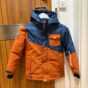 Blå och orange vinterjacka Cool Clubb - Snygg vinterjacka från Cool Club i blått och orange med huva, dragkedja och flera fickor. Jackan har fleecefoder, snölås och vattentätt material. Perfekt för kalla dagar och vinteraktiviteter. Märke på ärmen och smarta detaljer som justerbara ärmslut.