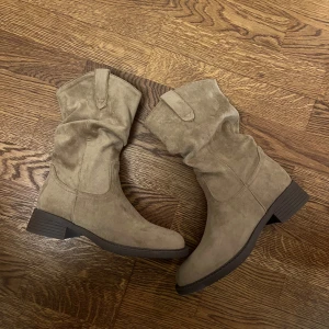 Beiga mockaboots med låg klack - Snygga boots i beige mocka med låg klack och rund tå. Skorna har en avslappnad, lite slouchy passform och bredare skaft med dekorativa sömmar och dragflikar upptill. Perfekta för dig som gillar en stilren och trendig look.