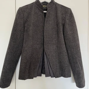 Grå rutig kavaj från LaVista Fashion - Snygg grå kavaj med diskret rutig struktur från LaVista Fashion. Jackan har lång ärm, rak passform och är fodrad med slät insida. Materialet är en blandning av ull och syntet, med detaljer i skinnimitation längs insidan. Perfekt för dig som gillar stilrena och klassiska plagg.