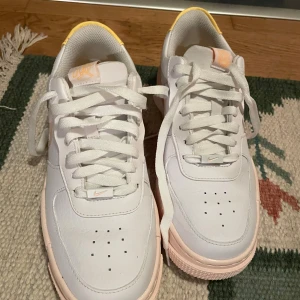 Nike Air Force 1 vit/rosa/gul sneakers - Nike Air Force 1 sneakers i vitt skinn med ljusrosa detaljer på sula och swoosh samt gul hälpanel. Klassisk rund tå, snörning och platt sula. Fräscha färgkombinationer och snygg design som sticker ut. Perfekt för dig som gillar streetstyle och vill ha något unikt.