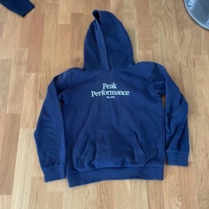 Blå hoodie Peak Performance 160 - Mörkblå hoodie från Peak Performance med stor vit logga på bröstet. Klassisk känguruficka framtill, mjuk insida och huva. Perfekt för dig som gillar en enkel och stilren look. Passar både tjejer och killar.