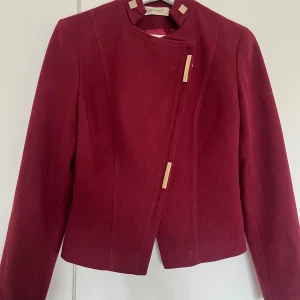 Vinröd kavaj från Garment Fashion - Snygg vinröd kavaj med guldiga detaljer och kedja bak från Garment Fashion. Jackan har asymmetrisk dragkedja framtill, långärmad design och coola fyrkantiga knappar vid kragen. Materialet är mjukt och känns lyxigt, perfekt för dig som gillar statement-plagg.