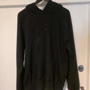 Svart hoodie från Giuseppe Zanotti - Svart hoodie från Giuseppe Zanotti med glittrande svart logga framtill. Klassisk känguruficka, huva och långa ärmar. Mjuk insida och relaxed fit, perfekt för en chill och snygg look.