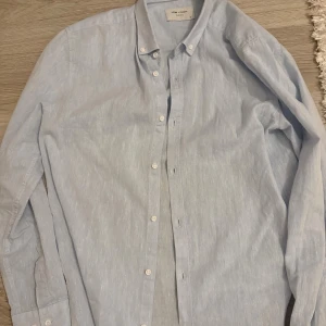 Ljusblå skjorta  - Säljer en stilren ljusblå skjorta. Skjortan har klassisk krage, knäppning framtill och långa ärmar. Perfekt för dig som gillar en clean och enkel look. Tillverkad i ett lätt och svalt material.