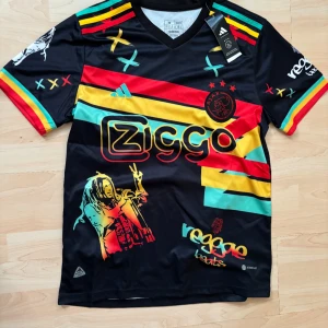Ajax x Bob Marley kortärmad fotbollströja - Ajax special edition fotbollströja från Adidas med Ziggo-logga, Bob Marley-silhuett och färgglada reggae-inspirerade ränder i rött, gult och grönt. Svart bas, korta ärmar och tryckta detaljer. Materialet är lätt och andas, perfekt för match eller träning.