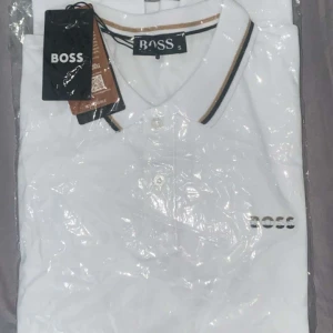 Vit pikétröja från BOSS med logga - Snygg vit pikétröja från BOSS med klassisk krage och knäppning framtill. Tröjan har diskret BOSS-logga på bröstet och stilrena ränder i svart och beige på kragen. Perfekt för en clean och fräsch look.