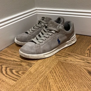 Grå mocka sneakers från  Ralph Lauren - Snygga grå sneakers i mocka från Polo Ralph Lauren med klassisk blå logga på sidan.Perfekt till jeans. De har tyvärr hål på insidan av  hälen där uppe se på bild går och fixa lätt med en lag lappar.