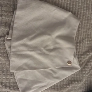 Vit omlottkjol med knappdetalj - Snygg vit omlottkjol med en stilren guldfärgad knappdetalj framtill. Kjolen har en kort och modern passform som ger en fräsch look. Perfekt för dig som gillar minimalistisk stil och vill ha något som sticker ut med en enkel detalj.