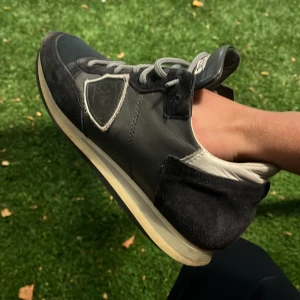 Svarta sneakers från Philippe Model - Säljer ett par svarta Philippe Model sneakers med grå detaljer och vit sula. Skorna har snörning, rund tå och är tillverkade i en mix av skinn och mocka. Klassisk sköldlogga på sidan och kontrasterande vit häl. Perfekta för dig som gillar stilrena och sportiga vibes.