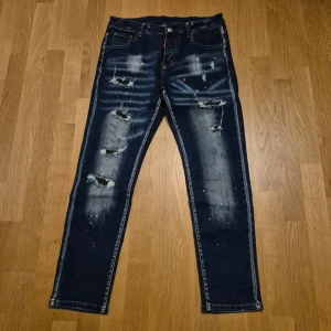 Dsquared2 slim jeans med slitningar - Mörkblå slim fit jeans från Dsquared2 med slitna detaljer, färgstänk och blekta partier. Klassisk femficksmodell med knappgylf och Dsquared2-logga bak i midjan. Snygg streetstil med unik finish, tillverkade i Italien.