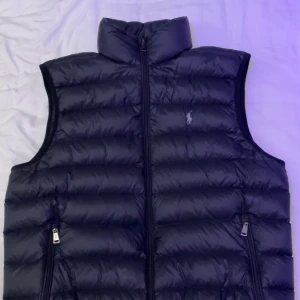 Svart dunväst från Polo Ralph Lauren - Snygg svart dunväst från Polo Ralph Lauren med horisontella quiltade paneler och hög krage. Västen har två fickor med dragkedja och en diskret logga på bröstet. Materialet är glansig polyester och dunfyllning, perfekt för lager-på-lager under kyliga dagar.