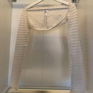 Vit bolerotopp med spetsdetaljer - Säljer en vit bolerotopp med lång ärm och spetsliknande struktur. Toppen är stickad i ett luftigt mönster och har en croppad passform som sitter snyggt över axlarna. Perfekt att slänga över ett linne eller klänning för en trendig layering-effekt.