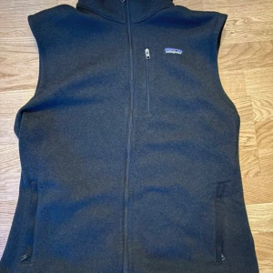 Mörkgrå stickad väst från Patagonia - Mörkgrå stickad väst från Patagonia med hel dragkedja framtill och två sidofickor med dragkedja. Västen har en hög krage och en liten bröstficka med dragkedja. Perfekt för lager-på-lager och outdoor vibes.