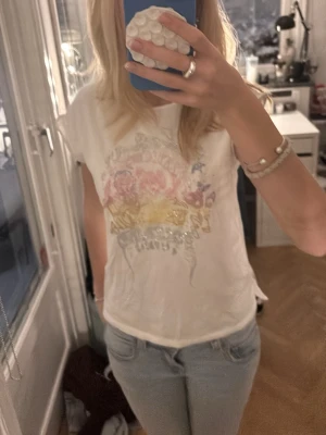 Vit t-shirt från Zadig & Voltaire - Säljer en vit t-shirt från Zadig & Voltaire med färgglatt tryck av blommor, döskalle och coola strassdetaljer i flera färger. T-shirten har korta ärmar och en avslappnad passform. Perfekt för dig som gillar statement-plagg och vill sticka ut lite extra.
