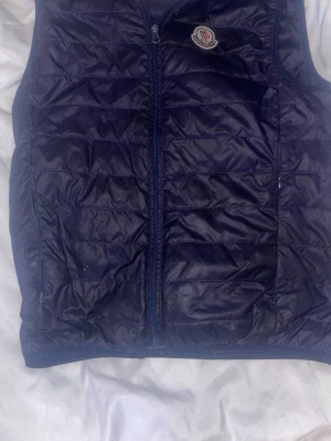 Mörkblå dunväst från Moncler - Säljer en mörkblå dunväst från Moncler med quiltad design och dragkedja framtill. Västen har ett klassiskt Moncler-märke på bröstet och är tillverkad i glansig polyester. Perfekt att slänga över en hoodie för extra värme under höst och varmare vinter. OBS dragkedjan är sönder och behöver bytas men har inte stört mig speciellt mycket!
