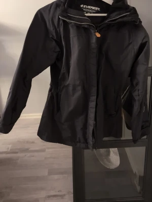 Svart vindjacka från Everest - Svart vindjacka från Everest med huva och dragkedja framtill. Jackan har två fickor med lock och en snygg, sportig look. Materialet är slitstark polyester och modellen är normal passform med långa ärmar. Perfekt för blåsiga dagar och outdoor.