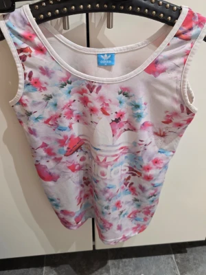 Blommig linnetopp från Adidas - Säljer en snygg linnetopp från Adidas i storlek M. Toppen är vit i grunden med ett färgglatt blommigt mönster i rosa, blått och rött. Adidas-loggan är tryckt framtill. Perfekt för sommardagar och chill outfits.