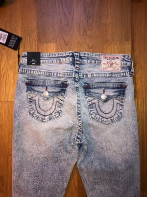 True Religion ljusblå bootcut jeans - Säljer ett par ljusblå bootcut jeans från True Religion med slitna detaljer och röda sömmar på bakfickorna. Jeansen har klassisk femficksdesign, knappar och dragkedja framtill samt tydlig tvättad look. Materialet är jeans och passformen är normal.