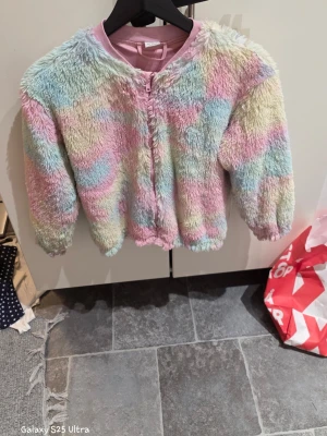 Pastellfärgad teddyjacka från Lindex - Säljer en fluffig teddyjacka från Lindex i storlek 134/140. Jackan har ett mjukt pastellmönster i rosa, blått, gult och lila, med dragkedja framtill och ribbad krage. Materialet är 100% polyester och jackan har långärmade ärmar. Perfekt för dig som gillar färgglada och mysiga plagg!