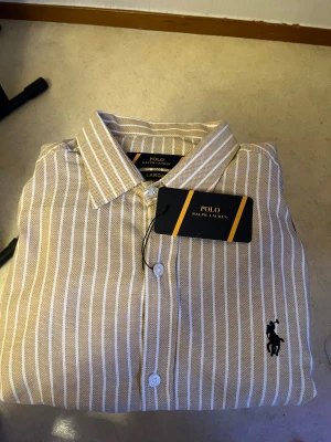 Beige randig skjorta från Ralph Lauren - Snygg beige skjorta från Ralph Lauren med vita vertikala ränder och klassisk krage. Skjortan har knappar framtill och den ikoniska broderade loggan i svart på bröstet. Perfekt för dig som gillar stilrena och tidlösa plagg.