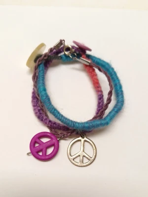 Färgglad armband med peace-berlocker - Unikt armbandi blått, lila och rosa med två stora peace-symboler framtill, en i metall och en i lila plast. Armbandet har en bohemisk vibe och är gjord i mjukt garn som ger en skön känsla. Perfekt för dig som vill sticka ut och addera färg till din outfit.