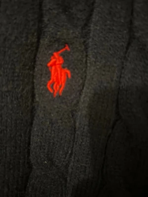 Svart stickad tröja Polo Ralph Lauren - Säljer en svart stickad tröja från Polo Ralph Lauren i storlek M. Tröjan har ett klassiskt kabelstickat mönster och en röd broderad logga på bröstet. Rund hals och långa ärmar, perfekt för dig som gillar stilren och tidlös design.