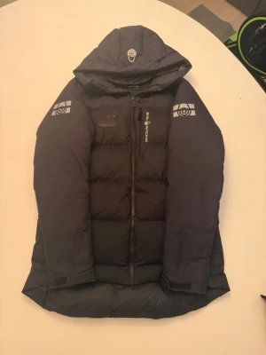  Sail Racing svart dunjacka  - Köpt på stadium förra året o använda inte längre, kvitto finns. Svart dunjacka från Sail Racing med huva och coola loggor på ärmar och bröst. Jackan har dragkedja framtill och är fodrad med 500 FP dun. Snygg, puffad design och detaljer som snörning i nederkant. Perfekt för kalla dagar och har en modern look.