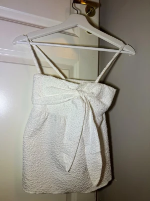 Vit omlott linnetopp med rosett - Säljer en vit linnetopp med smala axelband och snygg omlott-detalj framtill som knyts till en rosett. Toppen har en strukturerad, mönstrad yta och är lätt och luftig, perfekt för varma dagar. Passar dig som gillar en feminin och fräsch stil.