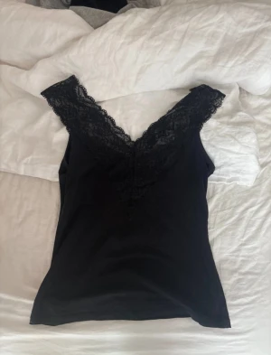 Svart linnetopp med spetsdetaljer - Snygg svart linnetopp med djup v-ringning och breda spetskanter längs halsringningen. Toppen är ärmlös och har en figurnära passform som framhäver siluetten. Perfekt att styla med jeans eller kjol för en trendig look.