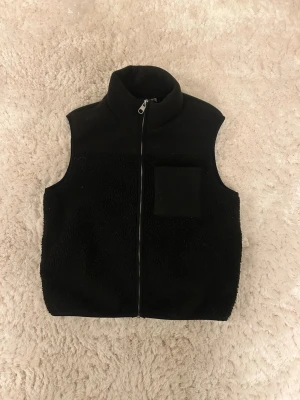 Svart teddyväst från Pieces XS - Svart teddyväst från Pieces i storlek XS. Västen har en hög krage, dragkedja framtill och en liten ficka på bröstet. Materialet är mjukt och fluffigt, perfekt för lager-på-lager. Enkel och stilren design som funkar till många outfits.