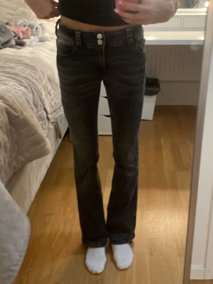 Svarta bootcut jeans med låg midja - Snygga svarta jeans med bootcut-snitt och låg midja. Jeansen har dubbla knappar fram, klassiska fickor och en cool tvättad look. Materialet är jeans och de har en figurnära passform upptill som blir vidare nedtill. Perfekta för dig som gillar en trendig siluett.