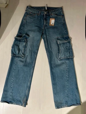 Blå cargojeans med stora fickor - Säljer ett par blå cargojeans med raka ben och stora sidofickor. Jeansen har klassisk femficksdesign, bälteshällor och är tillverkade i slitstarkt denim. Perfekta för dig som gillar en avslappnad och trendig stil.