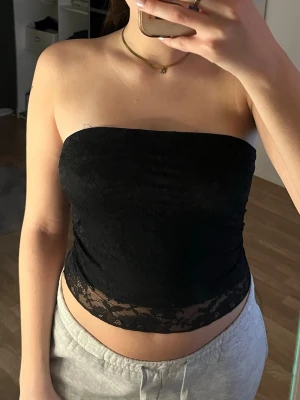 Svart spets tubtopp - Säljer en svart tubtopp med blommig spets och transparent nederkant. Toppen är axelbandslös och har en tight passform.