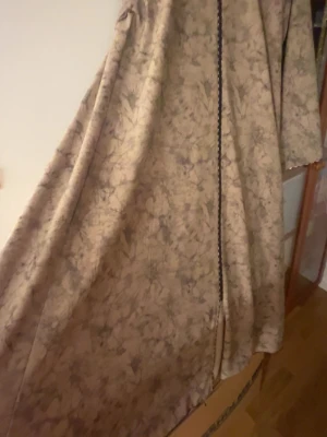 Beige lång klänning med dragkedja  - Säljer en beige lång klänning med grått marmorerat mönster och hel dragkedja framtill. Klänningen har ståkrage, vida långa ärmar och ett flowy fall. Perfekt för dig som vill ha en unik och stilren look. Den är väldigt vid och ganska lång. Pris går alltid att diskuteras💕