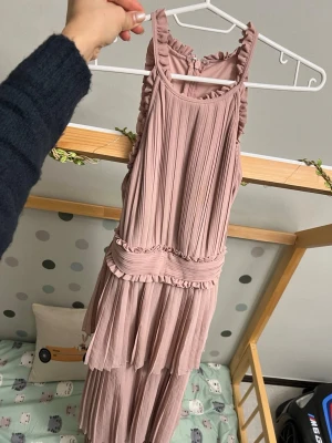 Ljusrosa plisserad långklänning - Supersöt ljusrosa långklänning med plisserat tyg och volangdetaljer längs kanterna och midjan. Klänningen har ärmlös design och rundad halsringning, perfekt för dig som gillar romantisk och flowy stil.