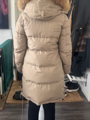 Beige dunjacka Parajumpers M - Säljer en beige Parajumpers dunjacka med huva och äkta päls, perfekt för kalla dagar. Jackan har dragkedja, snörning nedtill och praktiska fickor. Lång modell med quiltad design och gul detalj vid halsen. Slim fit och riktigt varm. Endast använd fåtal gånger så ser så gott som ny ut + den är kemtvättad. Finns både orginal kvitto från köp av jacka och från kemtvätt! 