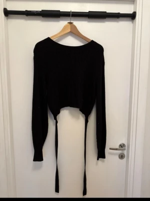 Svart croppad stickad tröja med knyt - Säljer en svart croppad stickad tröja med långa ärmar och snygg knytning baktill. Tröjan har en rund halsringning och är gjord i ett grovstickat material som känns mjukt och skönt. Perfekt för dig som gillar trendiga detaljer och vill ha en cool look.