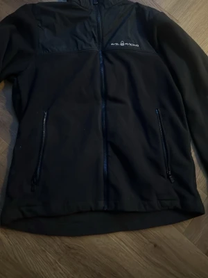 Svart fleecejacka från Sail Racing - Svart fleecejacka från Sail Racing med dragkedja framtill och två sidofickor med dragkedja. Jackan har broderad logga på bröstet och är tillverkad i mjuk fleece med slät yta upptill. Perfekt för kyliga dagar och har en normal passform.