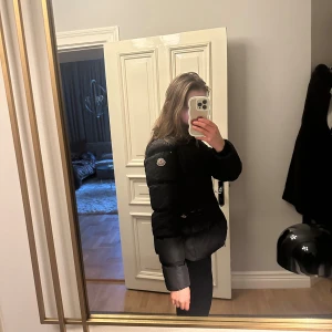 Svart dunjacka från Moncler - Snygg svart dunjacka från Moncler med markerad midja och dragkedja framtill. Jackan har två fickor med dragkedja och Moncler-logga på ärmen. 