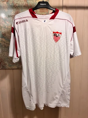Sevilla FC Joma vit/röd home shirt, storlek 3XL - Barely used.  #joma #sevillafc #laliga #vintage #spain #kanouté #fabiano #danialves #christianpoulsen   Vit Sevilla FC fotbollströja med röda detaljer. Tröjan har klubbens emblem på bröstet och La Liga-märket på ärmen. Den är dekorerad med subtila tryck av ”SEVILLA F.C.” och spelarnamn över hela tröjan.