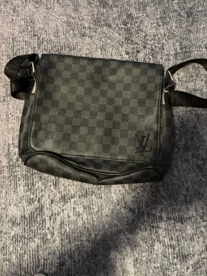 Louis Vuitton axelväska  - Väskan är svart med rutigt mönster och har justerbar axelrem med LV-logga på framsidan. Tillverkad i slitstarkt skinn och textil, perfekt för dig som vill ha en stilren och lyxig väska.